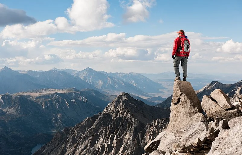 Ein Mann steht auf einer Felsspitze und blickt auf eine unter ihm liegende karge Gebirgslandschaft.