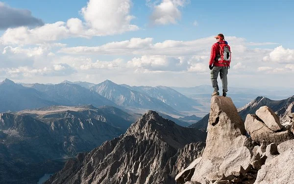 Ein Mann steht auf einer Felsspitze und blickt auf eine unter ihm liegende karge Gebirgslandschaft.