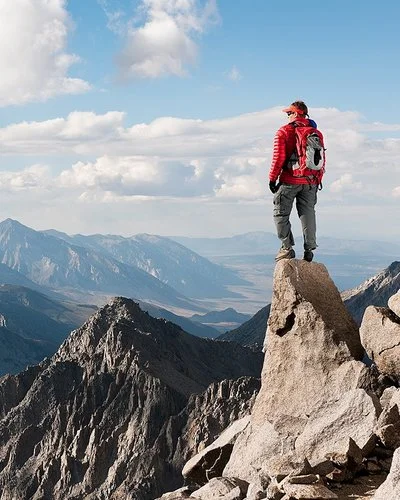 Ein Mann steht auf einer Felsspitze und blickt auf eine unter ihm liegende karge Gebirgslandschaft.