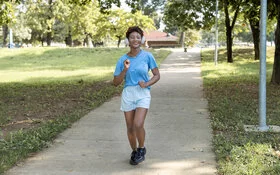 Frau macht Slow Jogging in einem Park, sie trägt dabei Kopfhörer auf ihren Ohren und ein Lächeln im Gesicht.