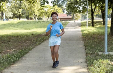 Frau macht Slow Jogging in einem Park, sie trägt dabei Kopfhörer auf ihren Ohren und ein Lächeln im Gesicht.