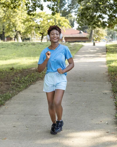 Frau macht Slow Jogging in einem Park, sie trägt dabei Kopfhörer auf ihren Ohren und ein Lächeln im Gesicht.