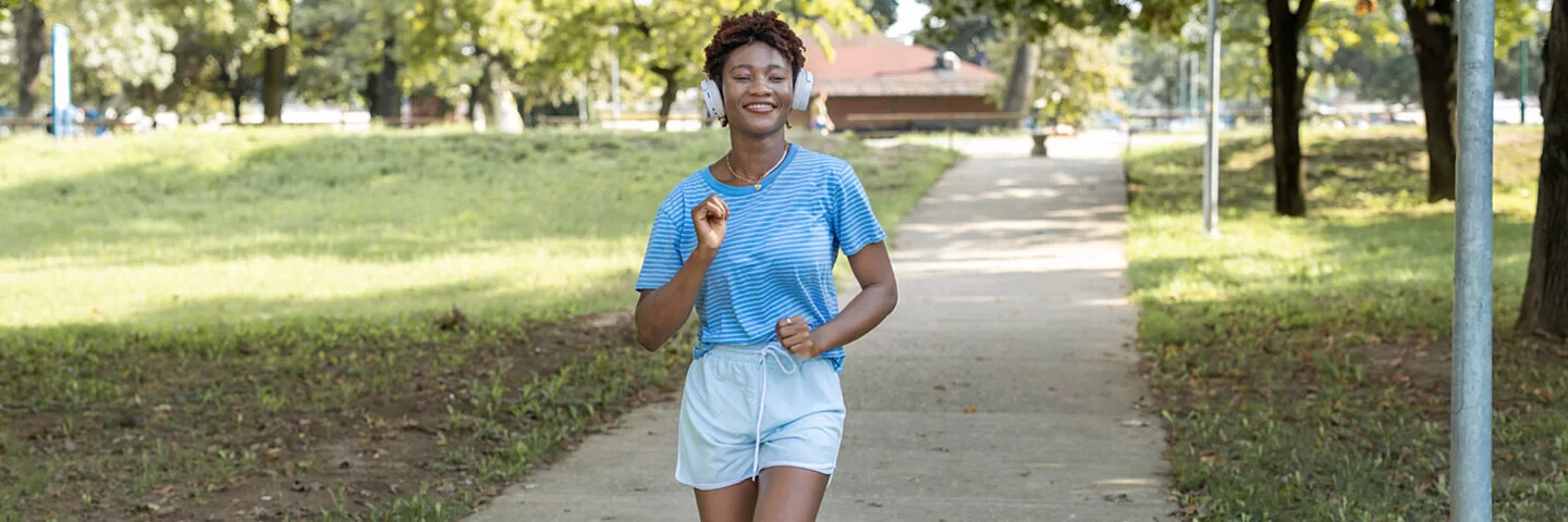Frau macht Slow Jogging in einem Park, sie trägt dabei Kopfhörer auf ihren Ohren und ein Lächeln im Gesicht.
