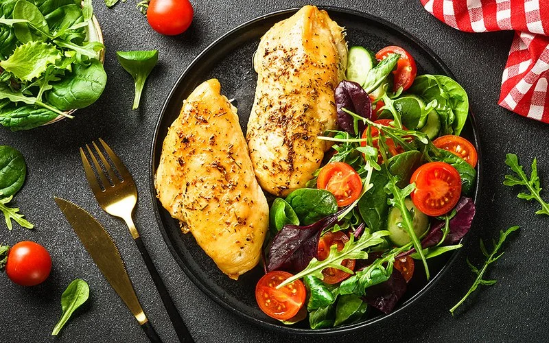 Ein frischer grüner Salat ist mit zwei Hähnchenfilets auf einem dunklen Teller angerichtet.