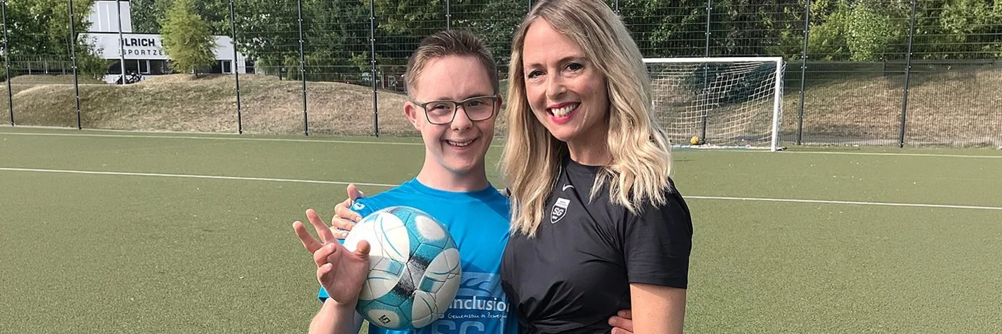 Ein Junge mit Down-Syndrom hält einen Fußball und steht lächelnd mit seiner Trainerin auf einem Fußballplatz.
