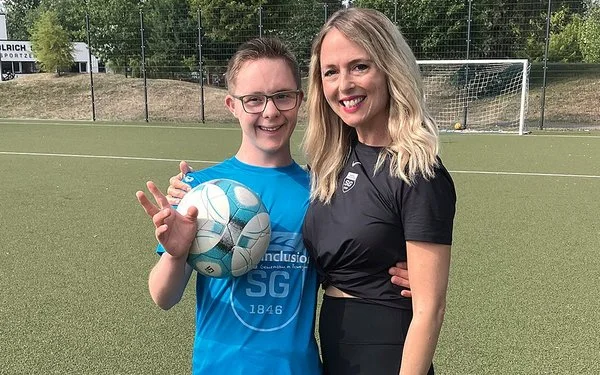 Ein Junge mit Down-Syndrom hält einen Fußball und steht lächelnd mit seiner Trainerin auf einem Fußballplatz.