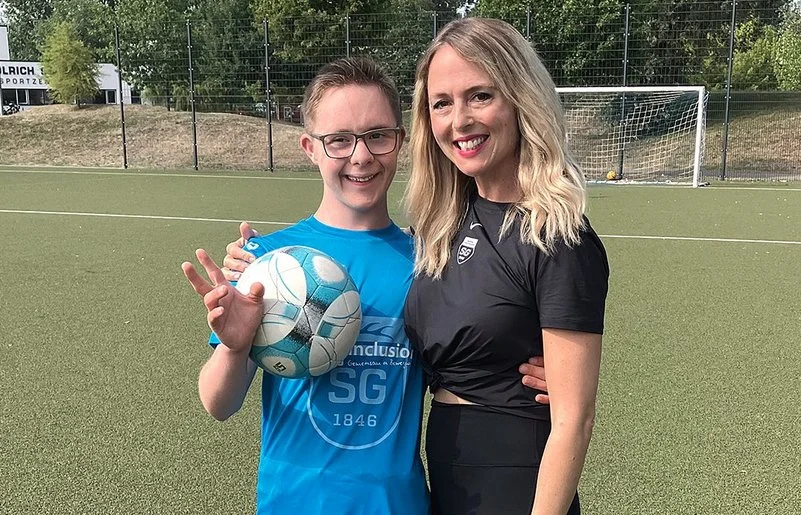 Ein Junge mit Down-Syndrom hält einen Fußball und steht lächelnd mit seiner Trainerin auf einem Fußballplatz.