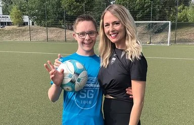 Ein Junge mit Down-Syndrom hält einen Fußball und steht lächelnd mit seiner Trainerin auf einem Fußballplatz.