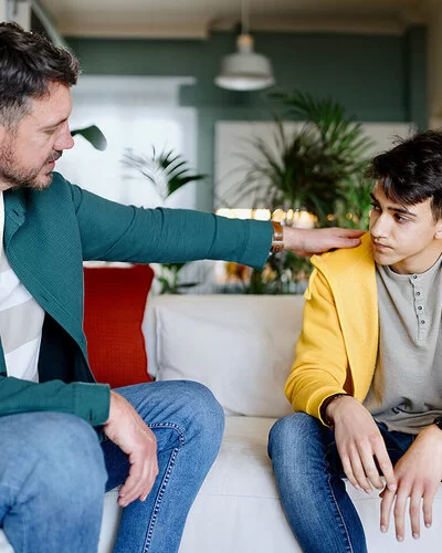 Ein besorgter Vater sitzt mit seinem jugendlichen Sohn auf der Couch und legt ihm die Hand auf die Schulter.