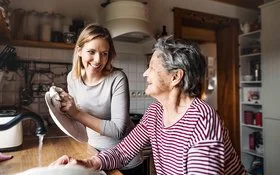 Eine junge und eine ältere Frau stehen gemeinsam in der Küche an einer Spüle. Sie unterhalten sich lachend, während die jüngere Frau das Geschirr abtrocknet. Im Hintergrund sind verschiedene Küchenmöbel, Regale und Küchenutensilien zu sehen.