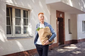 Ein junger Mann läuft eine Straße entlang. In der Hand hält er eine große Einkaufstüte aus Papier, die mit Lebensmitteln gefüllt ist.