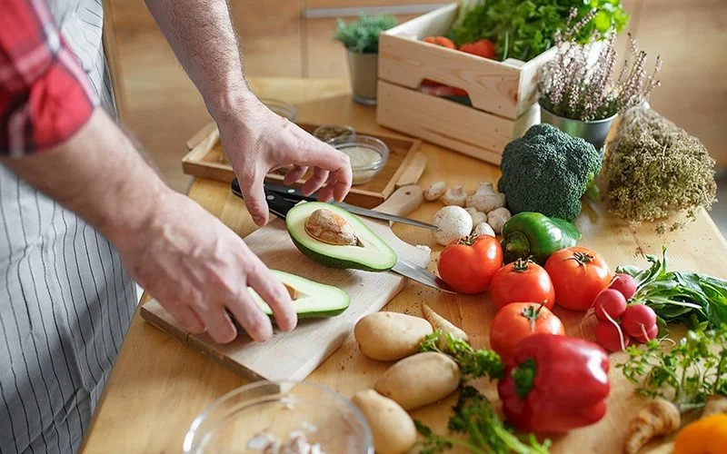 Ein Mann schneidet eine Avocado auf einem Holzbrett. Auf dem Tisch liegen verschiedene frische Gemüsesorten, darunter Tomaten, Kartoffeln, Brokkoli, Pilze und Kräuter. Im Hintergrund steht eine Holzkiste mit Salat und weiterem Gemüse.