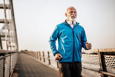 Ein sportlicher Senior bekleidet mit einer blauen Jacke läuft über eine Brücke.
