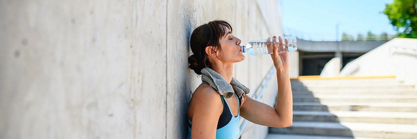 Eine junge Frau lehnt in Sportkleidung an einer grauen Betonwand und trinkt Wasser aus einer durchsichtigen Flasche.