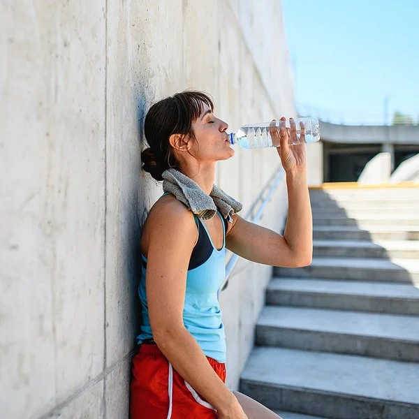 Eine junge Frau lehnt in Sportkleidung an einer grauen Betonwand und trinkt Wasser aus einer durchsichtigen Flasche.
