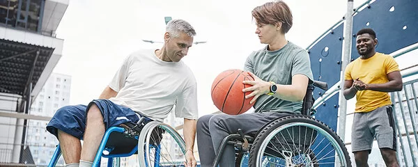 Eine Frau und ein Mann in Sport-Rollstühlen spielen Basketball auf einem Platz im Freien, ein afroamerikanischer Trainer steht im Hintergrund.