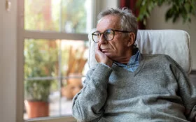 Ein älterer Mann mit Prostatakrebs steht vor dem Fenster und blickt besorgt in die Ferne.