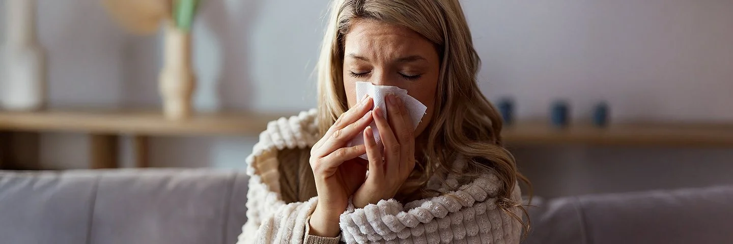 Eine Frau sitzt in eine Decke gehüllt auf einem Sofa und schnäutzt sich die Nase mit einem Papiertaschentuch.
