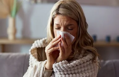 Eine Frau sitzt in eine Decke gehüllt auf einem Sofa und schnäutzt sich die Nase mit einem Papiertaschentuch.