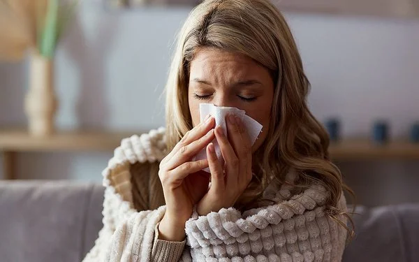 Eine Frau sitzt in eine Decke gehüllt auf einem Sofa und schnäutzt sich die Nase mit einem Papiertaschentuch.