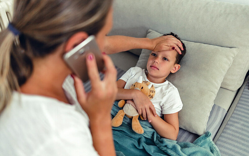 Eine Mutter fühlt die Temperatur an der Stirn ihres kranken Kindes und telefoniert mit einem Arzt.