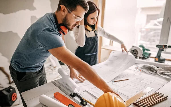 Eine Frau und ein Mann studieren Baupläne auf einer Haus-Baustelle, beide tragen geräuschisolierende Hörschützer um den Hals, der Mann trägt eine Schutzbrille.