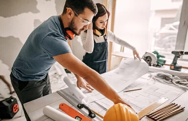 Eine Frau und ein Mann studieren Baupläne auf einer Haus-Baustelle, beide tragen geräuschisolierende Hörschützer um den Hals, der Mann trägt eine Schutzbrille.