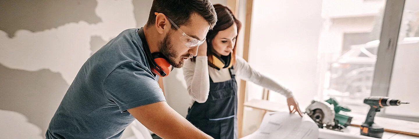 Eine Frau und ein Mann studieren Baupläne auf einer Haus-Baustelle, beide tragen geräuschisolierende Hörschützer um den Hals, der Mann trägt eine Schutzbrille.