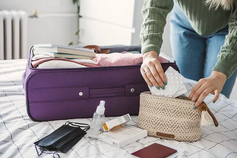 Eine Frau packt einen Koffer. Dieser liegt auf ihrem Bett. Zusätzlich packt sie eine kleine Kosmetiktasche, in der sie Medikamente verstaut.