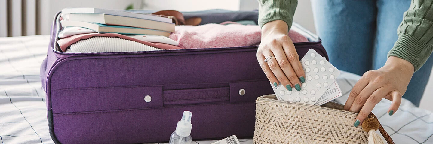 Eine Frau packt einen Koffer. Dieser liegt auf ihrem Bett. Zusätzlich packt sie eine kleine Kosmetiktasche, in der sie Medikamente verstaut.