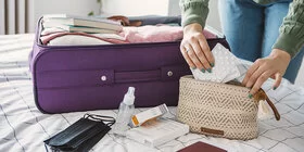 Eine Frau packt einen Koffer. Dieser liegt auf ihrem Bett. Zusätzlich packt sie eine kleine Kosmetiktasche, in der sie Medikamente verstaut.