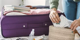 Eine Frau packt einen Koffer. Dieser liegt auf ihrem Bett. Zusätzlich packt sie eine kleine Kosmetiktasche, in der sie Medikamente verstaut.