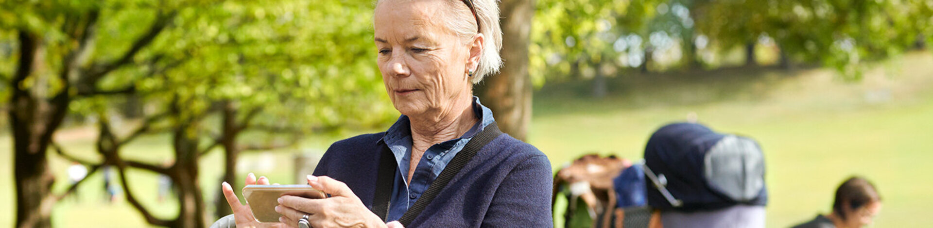 Eine ältere Dame sitzt im Park auf einer Bank und schaut sich Informationen auf ihrem Smartphone an.