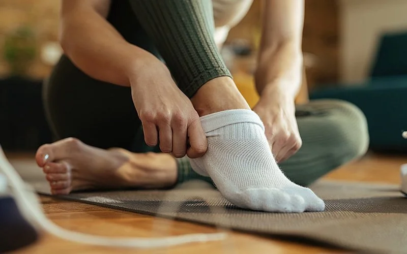 Eine Frau in sportlicher Kleidung auf einer Yoga Matte. Sie zieht sich eine weiße Socke über den rechten Fuß.