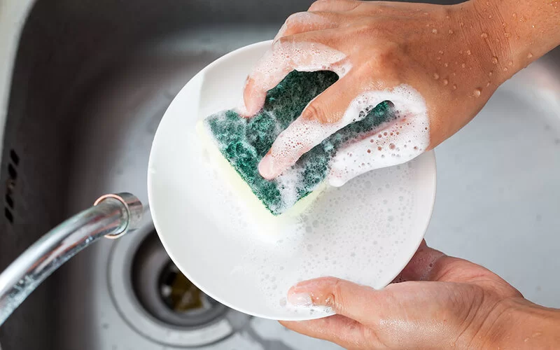 Die Hände einer Frau, die eine Untertasse mit einem Küchenschwamm in einer Spüle reinigt.