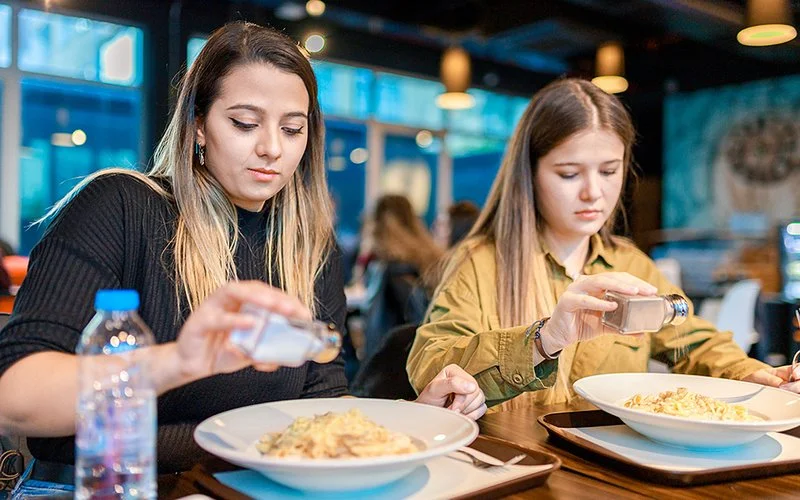 Eine Frau und ein Mädchen sitzen an einem Tisch und würzen ihr Essen mit Salz.