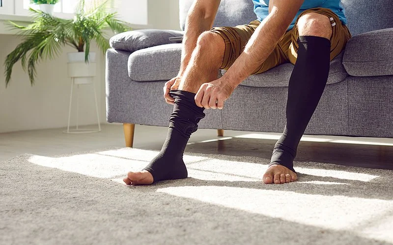 Ein Mann (zu sehen sind nur die Beine und die Arme) sitzt auf einem grauen Sofa und zieht sich schwarze Kompressionsstrümpfe an.