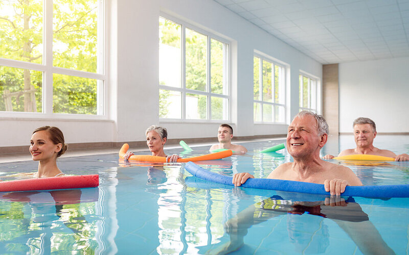 Eine Gruppe von Menschen unterschiedlichen Alters macht in einem Schwimmbecken Wassergymnastik mit Schwimmnudeln.
