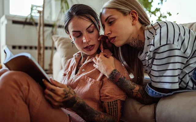 Zwei Frauen mit langen Haaren und Tätowierungen im Wohnzimmer. Eine Frau liegt auf der Couch und lehnt sich an die andere, die auf dem Boden sitzt und ein Buch liest, beide schauen ernst und etwas angespannt.