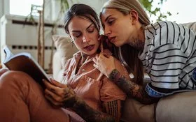 Zwei Frauen mit langen Haaren und Tätowierungen im Wohnzimmer. Eine Frau liegt auf der Couch und lehnt sich an die andere, die auf dem Boden sitzt und ein Buch liest, beide schauen ernst und etwas angespannt.