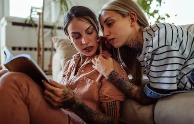 Zwei Frauen mit langen Haaren und Tätowierungen im Wohnzimmer. Eine Frau liegt auf der Couch und lehnt sich an die andere, die auf dem Boden sitzt und ein Buch liest, beide schauen ernst und etwas angespannt.