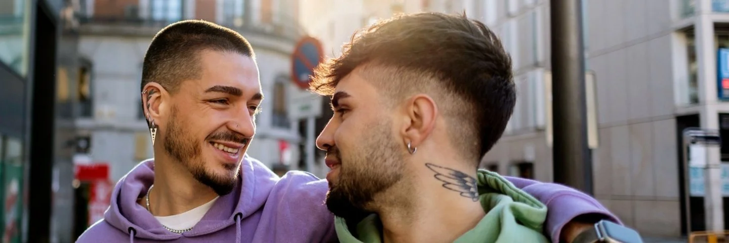 Zwei junge Männer mit Drei-Tage-Bart gehen Arm in Arm durch eine Innenstadt. Sie schauen sich dabei verliebt an.