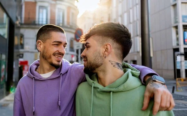 Zwei junge Männer mit Drei-Tage-Bart gehen Arm in Arm durch eine Innenstadt. Sie schauen sich dabei verliebt an.