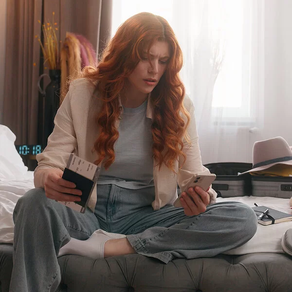 Reiseangst: Eine Frau sitzt auf ihrem Bett, um sie herum ist das Gepäck verstreut. Sie hält das Flugticket in der rechten und ihr Handy in der linken Hand.