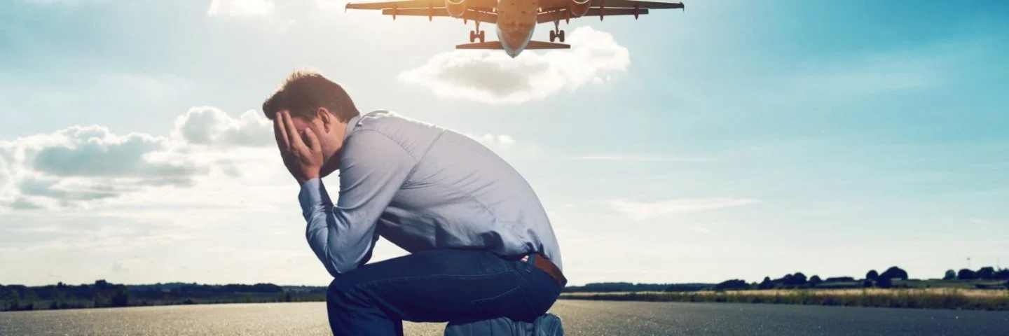 Ein Mann sitzt mit aufgestütztem Kopf auf einem Koffer auf dem Rollfeld eines Flughafens. Im Hintergrund ist ein aufsteigendes Flugzeug zu sehen.