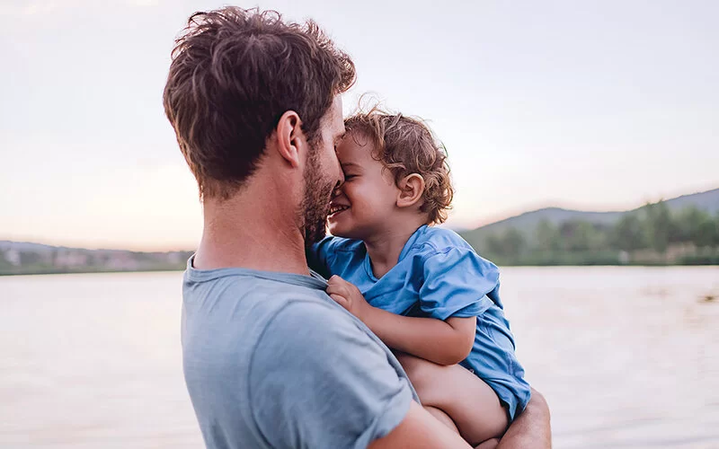 Ein Vater steht an einem See und trägt sein kleines Kind liebevoll auf dem Arm.