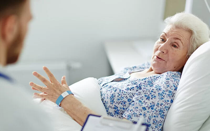 Eine ältere Patientin liegt in einem Krankenbett und spricht mit einem Arzt über die Vor- und Nachteile von Stents und Bypässen.
