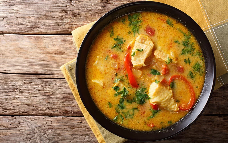 Auf einem Holztisch steht auf einer gelbkarierten Stoffserviette ein brasilianischer Paprika-Fisch-Suppe.