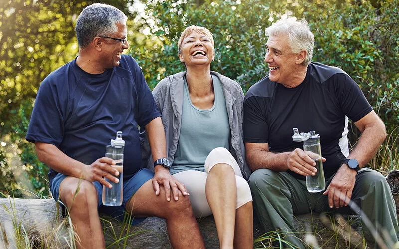 Drei ältere Menschen in Sportkleidung sitzen auf einem Baumstamm und lachen, zwei von ihnen trinken Wasser.