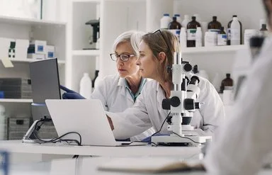 In einem medizinischen Labor sitzen zwei Frauen in weißen Kitteln an einem Arbeitstisch. Auf dem Tisch stehen ein Mikroskop, ein aufgeklappter Laptop und ein Monitor. Beide schauen auf den Monitor, auf dem eine der Frauen mit dem Zeigefinger auf etwas zeigt.